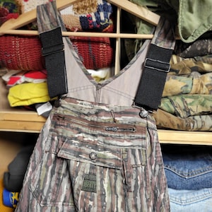 May include: Camouflage-patterned overalls with black straps and a zipper closure. The overalls have a pocket with a Liberty label. The garment is hanging on a wooden hanger in a closet with other clothing.