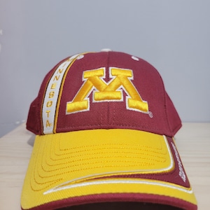 May include: A maroon and gold baseball cap featuring the embroidered yellow letter M on the front crown. A vertical white stripe on the left side displays the word MINNESOTA in gold text, and the curved brim is yellow with maroon trim.