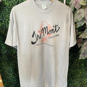 May include: A light gray short-sleeve t-shirt featuring a graphic with the text "Tri-Mont 1912-1988" in black script and block lettering, an orange line-art bird illustration, and the text "May the tale never be forgotten" printed below.