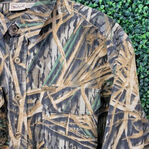 May include: A long-sleeved button-down shirt with a camouflage pattern in shades of brown, green, and gray. The shirt has two chest pockets and the Woolrich brand label is visible. The shirt is hanging on a wooden hanger.