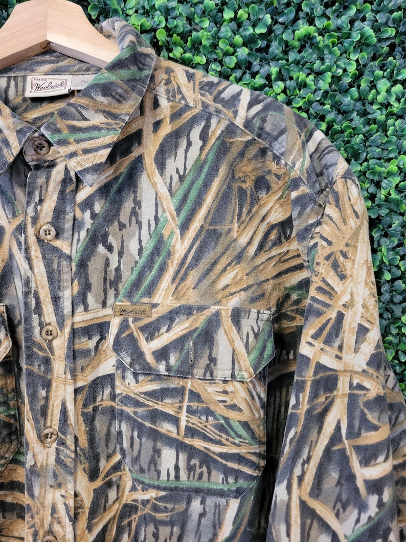 May include: A long-sleeved button-down shirt with a camouflage pattern in shades of brown, green, and gray. The shirt has two chest pockets and the Woolrich brand label is visible. The shirt is hanging on a wooden hanger.