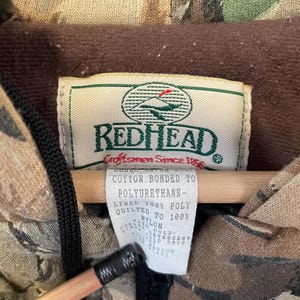 May include: Close-up of a camouflage jacket with a brown interior lining. The jacket has a tag that reads "REDHEAD Craftsmen Since 1856". The tag also lists the materials used, including cotton, polyurethane, and nylon. A black zipper is visible.