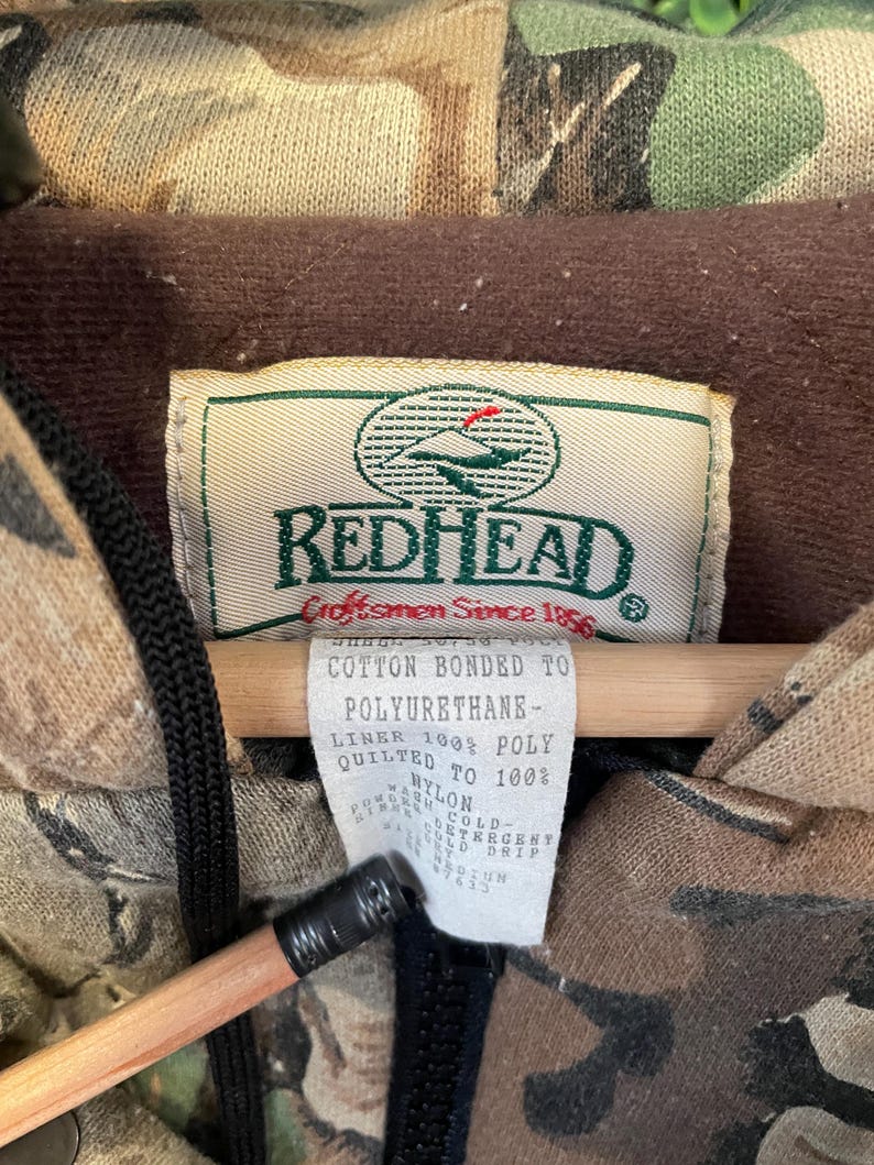 May include: Close-up of a camouflage jacket with a brown interior lining. The jacket has a tag that reads "REDHEAD Craftsmen Since 1856". The tag also lists the materials used, including cotton, polyurethane, and nylon. A black zipper is visible.