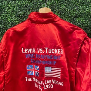May include: A vibrant red satin jacket with embroidered text and flags. The text reads "LEWIS VS. TUCKER WBC HEAVYWEIGHT CHAMPIONSHIP" with the flags of the United Kingdom and the United States. The text "THE MIRAGE, LAS VEGAS MAY 8, 1993" is also visible.