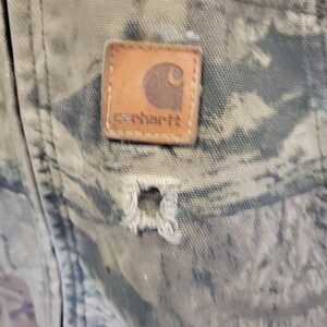 May include: Close-up of a camouflage-patterned fabric with a distressed appearance. A brown leather Carhartt brand patch is sewn onto the fabric. A hole is visible in the fabric below the patch.
