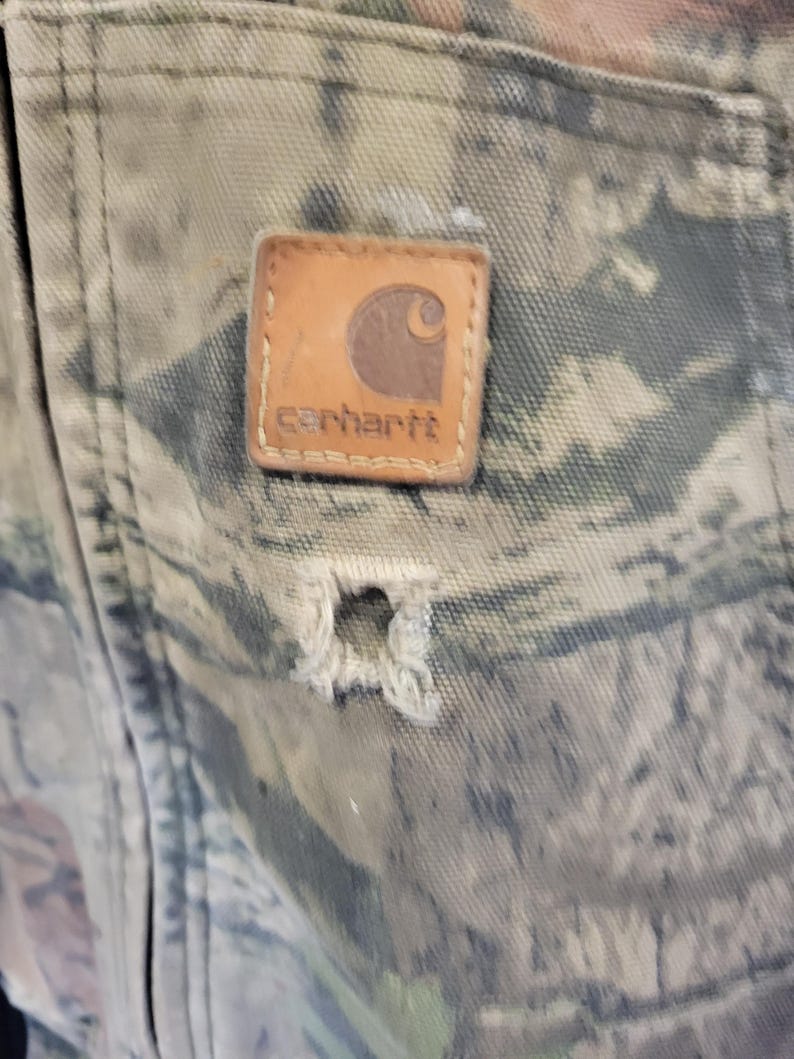 May include: Close-up of a camouflage-patterned fabric with a distressed appearance. A brown leather Carhartt brand patch is sewn onto the fabric. A hole is visible in the fabric below the patch.