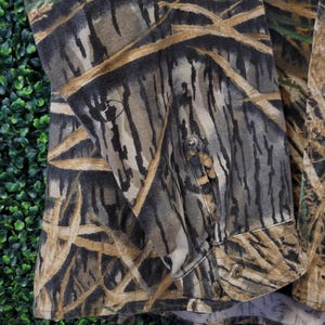May include: Camouflage button-down shirt in shades of brown, green, and tan. The fabric features a detailed pattern resembling a forest or wetland environment. The shirt has buttoned cuffs and a classic collar.