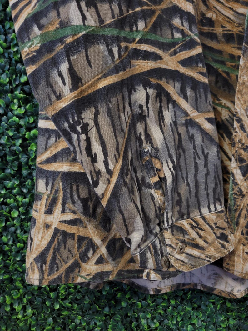 May include: Camouflage button-down shirt in shades of brown, green, and tan. The fabric features a detailed pattern resembling a forest or wetland environment. The shirt has buttoned cuffs and a classic collar.