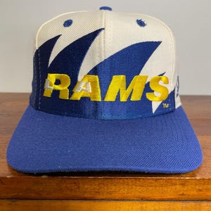 May include: A baseball cap featuring a cream-coloured crown and a blue brim. The front displays the word RAMS in bold yellow letters over a dark blue graphic design. The cap includes a small TM symbol near the right side of the lettering.