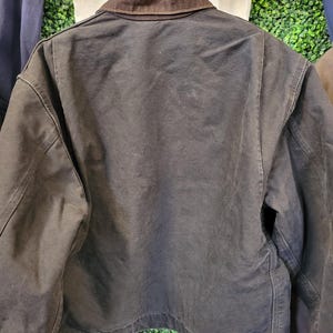 May include: A dark brown canvas jacket featuring a contrasting dark brown corduroy collar, long sleeves, and a straight hemline. The jacket includes metal button closures at the cuffs and along the bottom hem, showcasing a sturdy, heavy-duty construction.