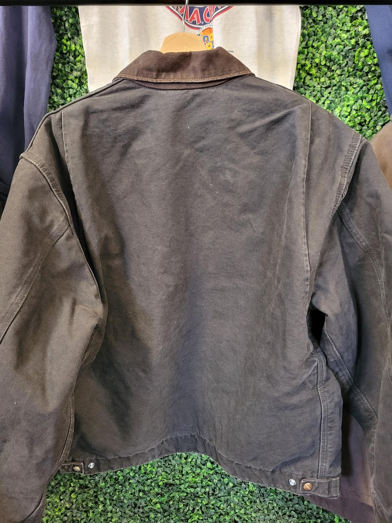 May include: A dark brown canvas jacket featuring a contrasting dark brown corduroy collar, long sleeves, and a straight hemline. The jacket includes metal button closures at the cuffs and along the bottom hem, showcasing a sturdy, heavy-duty construction.