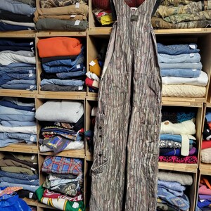 May include: Camouflage-patterned overalls hanging on a wooden hanger. The overalls have multiple pockets and button details. The background shows a wooden shelving unit filled with folded clothing items, including jeans, sweaters, and shirts.