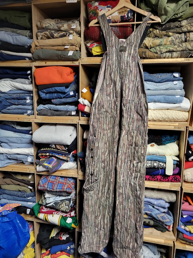 May include: Camouflage-patterned overalls hanging on a wooden hanger. The overalls have multiple pockets and button details. The background shows a wooden shelving unit filled with folded clothing items, including jeans, sweaters, and shirts.
