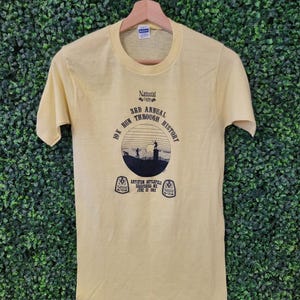 May include: A pale yellow t-shirt with a crew neck and short sleeves. The shirt displays a graphic with the text "Natural Light 10K Run Through History" and event details. The t-shirt is suspended on a wooden hanger against a green backdrop.