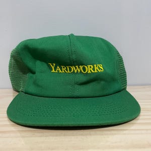 Może przedstawiać: Zielona czapka bejsbolowa z żółtym haftem "YARDWORKS". Czapka ma siatkowy tył i zakrzywiony daszek. Kapelusz jest wykonany z wytrzymałej tkaniny.