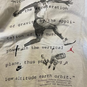 Pu&ograve; includere: T-shirt bianca con un'immagine in bianco e nero di Michael Jordan in azione, con testo che cita il professor Douglas Kirkpatrick. Sono visibili anche il logo Nike e il logo Jumpman.