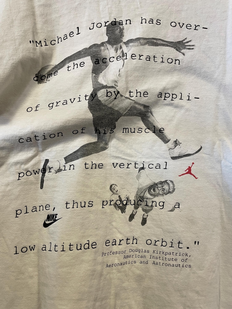 Pu&ograve; includere: T-shirt bianca con un'immagine in bianco e nero di Michael Jordan in azione, con testo che cita il professor Douglas Kirkpatrick. Sono visibili anche il logo Nike e il logo Jumpman.