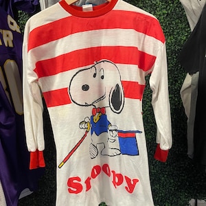 May include: A long-sleeve shirt featuring horizontal red and white stripes with a graphic of Snoopy dressed as a magician, wearing a blue jacket, red bowtie, and holding a red cane next to a blue top hat, with the red text Snoopy printed below.