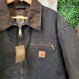 May include: A black Carhartt jacket with a brown corduroy collar, a gold-tone front zipper, and a chest pocket with a zipper. The jacket features a square Carhartt logo patch and a hanging tag that reads Carhartt Will Be Legendary in white text.
