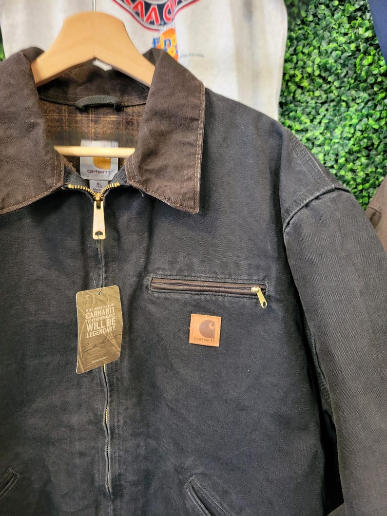 May include: A black Carhartt jacket with a brown corduroy collar, a gold-tone front zipper, and a chest pocket with a zipper. The jacket features a square Carhartt logo patch and a hanging tag that reads Carhartt Will Be Legendary in white text.