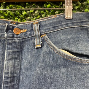 May include: Close-up of blue denim jeans with a button and pocket detail. The jeans have a classic design with visible stitching and a brass-coloured button. The fabric appears to be a medium wash denim.