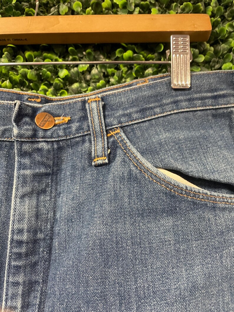 May include: Close-up of blue denim jeans with a button and pocket detail. The jeans have a classic design with visible stitching and a brass-coloured button. The fabric appears to be a medium wash denim.
