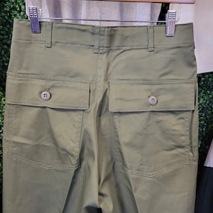 Op de afbeelding: Olijfgroene broek met twee achterzakken met knoopsluiting en een knoop- en ritssluiting. De broek hangt aan een houten kleerhanger. De stof lijkt een duurzaam katoen of mix te zijn.