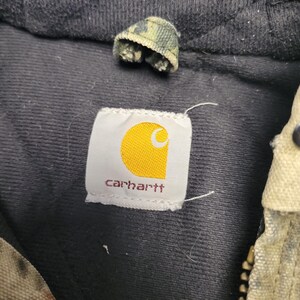 May include: Close-up of a Carhartt label with a yellow logo on a white background, and the brand name in red. The label is sewn onto a black fabric. A small camouflage fabric piece is visible above the label.