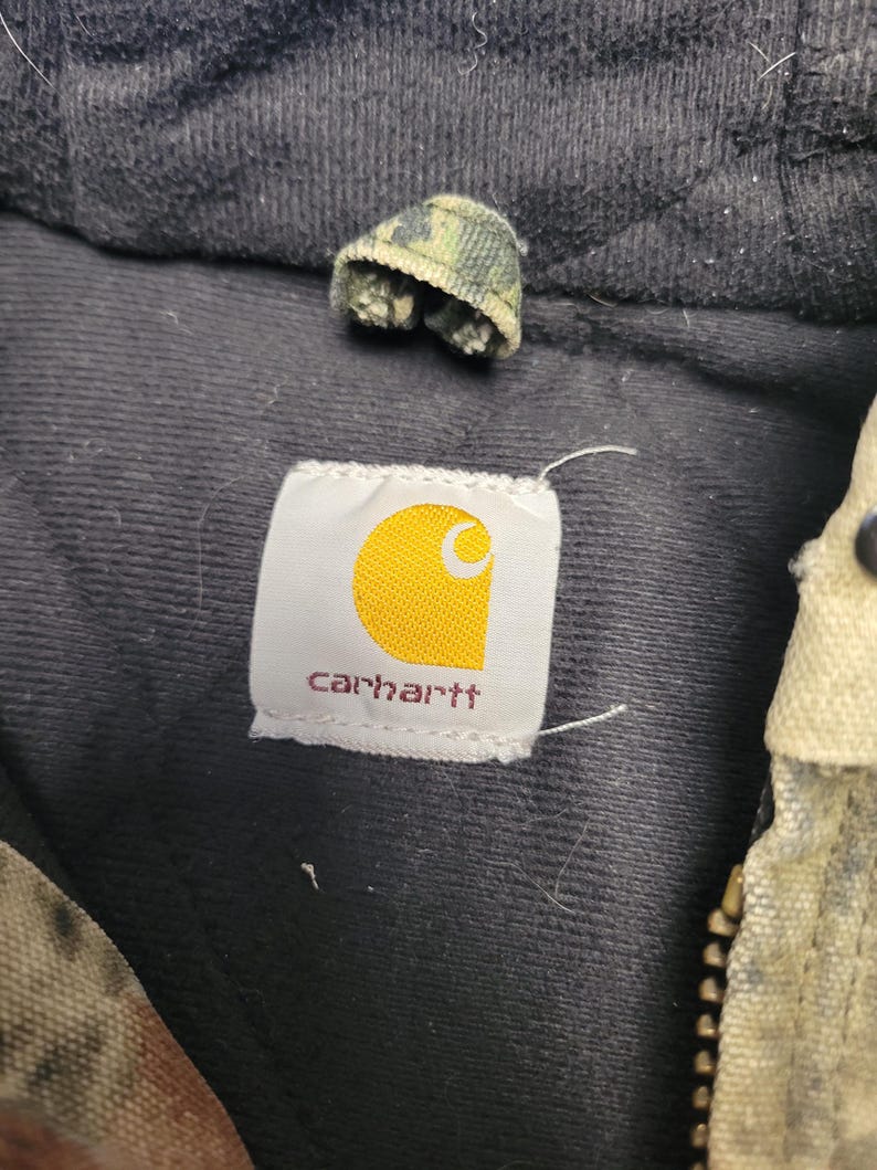May include: Close-up of a Carhartt label with a yellow logo on a white background, and the brand name in red. The label is sewn onto a black fabric. A small camouflage fabric piece is visible above the label.