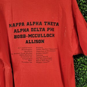 May include: Red t-shirt with black text. The text includes "KAPPA ALPHA THETA", "ALPHA DELTA PHI", "BOBB-MCCULLOCH", and "ALLISON". Additional text lists various names and places. The shirt is made of a soft material.