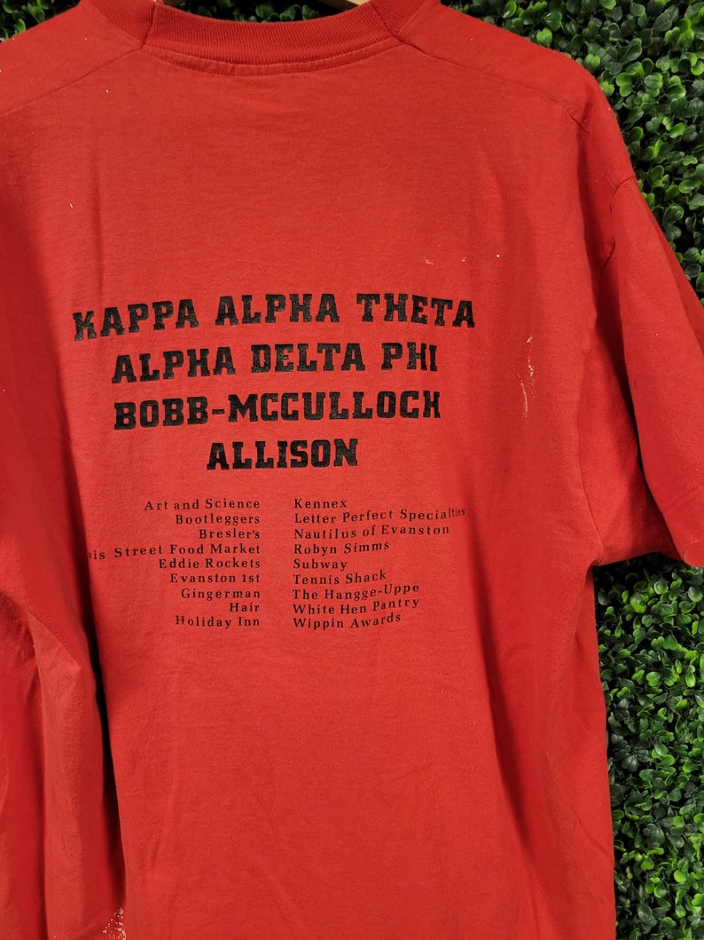 May include: Red t-shirt with black text. The text includes "KAPPA ALPHA THETA", "ALPHA DELTA PHI", "BOBB-MCCULLOCH", and "ALLISON". Additional text lists various names and places. The shirt is made of a soft material.