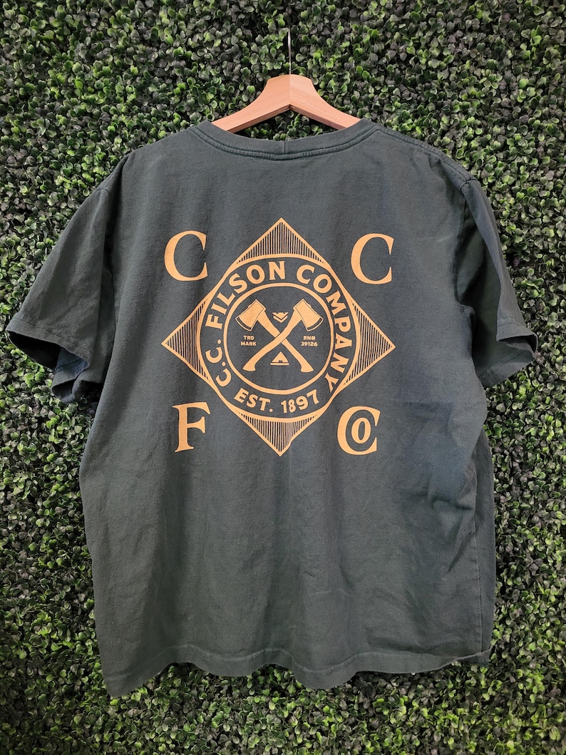 May include: Dark green t-shirt with a gold diamond-shaped graphic on the back. The graphic features crossed axes and the text "C.C. FILSON COMPANY EST. 1897". The shirt is hanging on a wooden hanger.