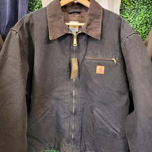 May include: A dark charcoal gray zip-up jacket featuring a brown corduroy collar, a gold-tone metal zipper, and a chest pocket with a zipper closure. A rectangular tan brand patch is visible on the chest, along with a hanging paper product tag.