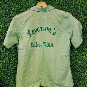 May include: Light green short-sleeved shirt with a collar. The back of the shirt features the embroidered text "Leverson's Odin, Minn." in a darker green script. The shirt is hanging on a wooden hanger against a green textured background.