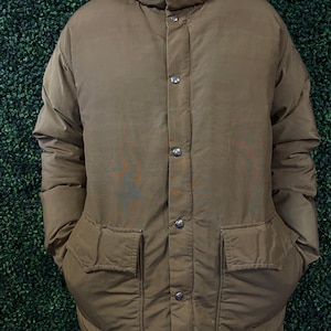 May include: A tan puffer jacket featuring a high collar, a front placket with silver-toned metal snap buttons, and two large patch pockets with flaps on the lower front. The jacket has a quilted construction and long sleeves with ribbed cuffs.