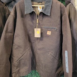 May include: A brown Carhartt jacket featuring a dark grey collar, a full-length brass zipper, and a plaid interior lining. The jacket includes a zippered chest pocket, two lower front pockets, and a Carhartt brand tag attached to the zipper.