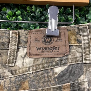 May include: Close-up of Wrangler Authentic Jeans with a camouflage pattern. The jeans have a brown leather patch with the Wrangler logo and text. The jeans are held by a silver metal clip attached to a wooden hanger.