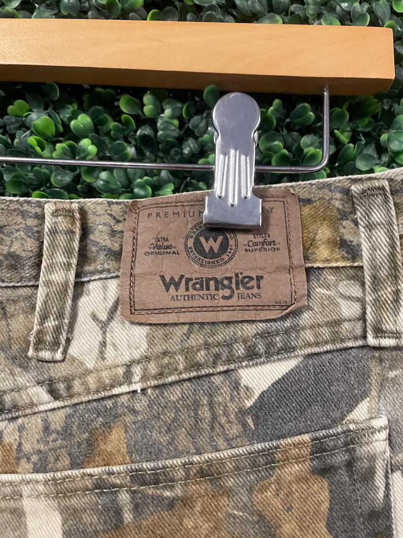 May include: Close-up of Wrangler Authentic Jeans with a camouflage pattern. The jeans have a brown leather patch with the Wrangler logo and text. The jeans are held by a silver metal clip attached to a wooden hanger.