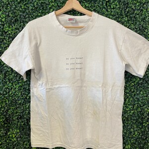 Pu&ograve; includere: Una maglietta bianca con la scritta "Do you know?" ripetuta tre volte in un font semplice. La maglietta &egrave; appesa a una gruccia di legno su uno sfondo verde. Il logo Nike &egrave; visibile sul colletto.