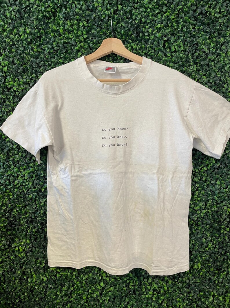Pu&ograve; includere: Una maglietta bianca con la scritta "Do you know?" ripetuta tre volte in un font semplice. La maglietta &egrave; appesa a una gruccia di legno su uno sfondo verde. Il logo Nike &egrave; visibile sul colletto.