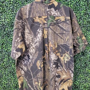Op de afbeelding: Korte mouwen camouflage shirt in tinten bruin, groen en beige, hangend aan een houten hanger. Het shirt heeft een knoopsluiting aan de voorkant en een klassieke kraag. De stof is bedrukt met een realistisch bosontwerp.