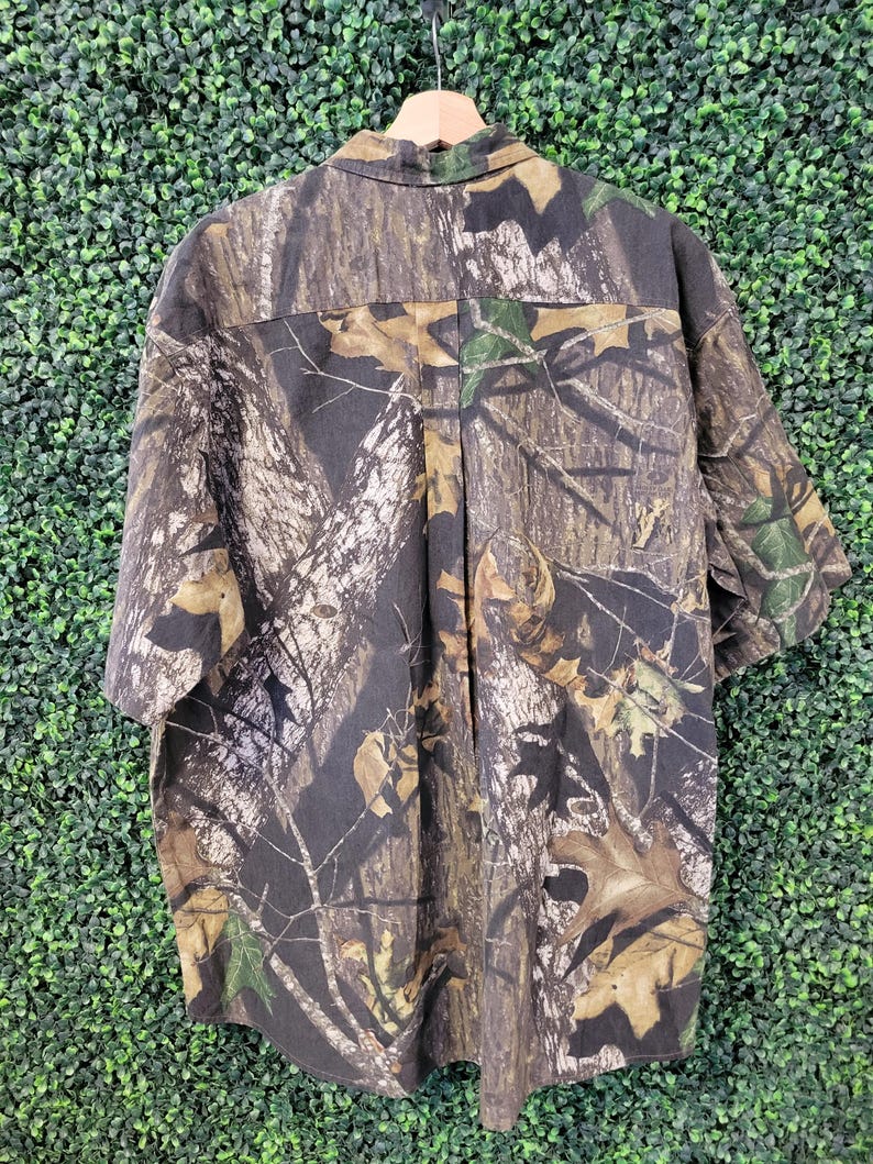 Op de afbeelding: Korte mouwen camouflage shirt in tinten bruin, groen en beige, hangend aan een houten hanger. Het shirt heeft een knoopsluiting aan de voorkant en een klassieke kraag. De stof is bedrukt met een realistisch bosontwerp.