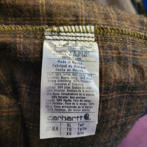 May include: A white rectangular garment care label stitched into a brown and black plaid flannel lining. The label features the Carhartt logo, size XL Tall, and text detailing material composition in English, French, and Spanish, including RN 14806.