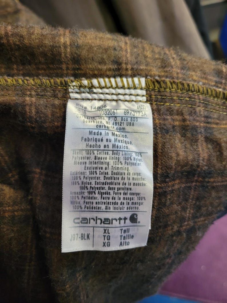 May include: A white rectangular garment care label stitched into a brown and black plaid flannel lining. The label features the Carhartt logo, size XL Tall, and text detailing material composition in English, French, and Spanish, including RN 14806.