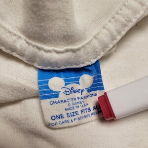 K&ouml;nnte beinhalten: Nahaufnahme eines wei&szlig;en Kleidungsst&uuml;cketiketts mit dem Disney-Logo und Text. Das Etikett ist blau und wei&szlig; mit den Worten "CHARACTER FASHIONS DISNEY MADE IN U.S.A. ONE SIZE FITS ALL FOR CARE & FIBER SEE REVER". Ein rot-wei&szlig;es Objekt befindet sich im Bild.