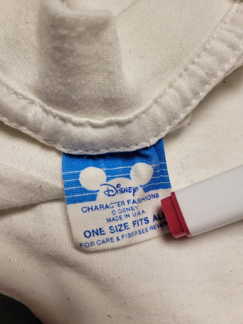 K&ouml;nnte beinhalten: Nahaufnahme eines wei&szlig;en Kleidungsst&uuml;cketiketts mit dem Disney-Logo und Text. Das Etikett ist blau und wei&szlig; mit den Worten "CHARACTER FASHIONS DISNEY MADE IN U.S.A. ONE SIZE FITS ALL FOR CARE & FIBER SEE REVER". Ein rot-wei&szlig;es Objekt befindet sich im Bild.