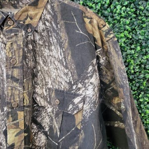 Op de afbeelding: Camouflage overhemd met knopen en twee borstzakken. Het overhemd heeft een patroon van bruin, groen en beige, dat lijkt op een bosrijke omgeving. Het overhemd is gemaakt van een duurzame stof en heeft een klassieke kraag.