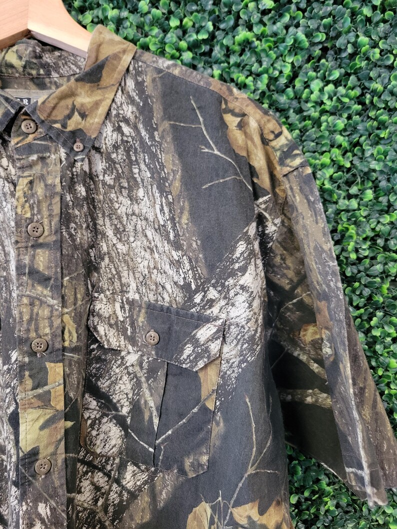 Op de afbeelding: Camouflage overhemd met knopen en twee borstzakken. Het overhemd heeft een patroon van bruin, groen en beige, dat lijkt op een bosrijke omgeving. Het overhemd is gemaakt van een duurzame stof en heeft een klassieke kraag.