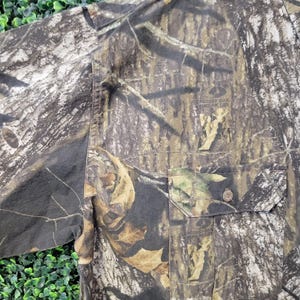 Op de afbeelding: Camouflage overhemd met knoopsluiting en een borstzak. Het overhemd heeft een patroon van bruin, groen en beige, met de tekst "Mossy Oak Break-Up" zichtbaar op de zak.