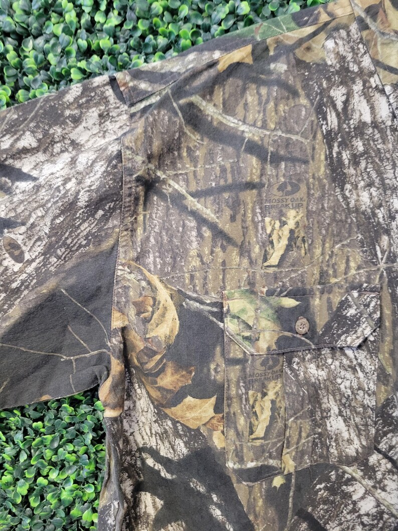 Op de afbeelding: Camouflage overhemd met knoopsluiting en een borstzak. Het overhemd heeft een patroon van bruin, groen en beige, met de tekst "Mossy Oak Break-Up" zichtbaar op de zak.